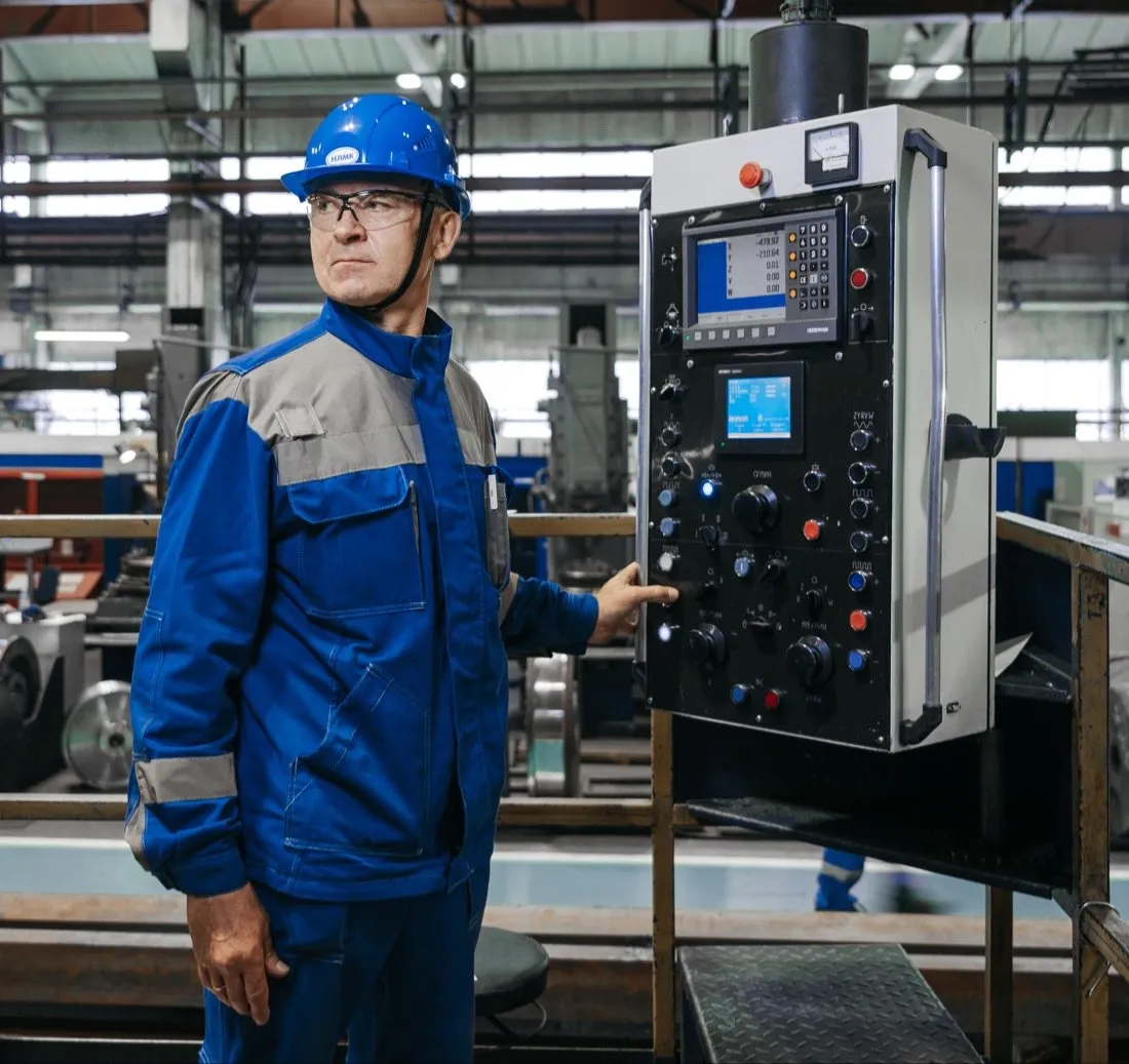 An engineer in a hard hat and safety glasses reviews machine data on a tablet on the plant floor